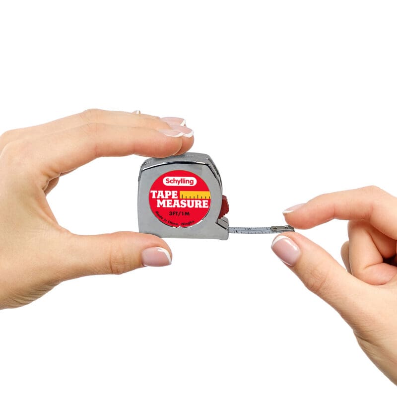 TPM_Image2 Two hands holding a miniature tape measure with a metallic body. The tape measure, branded "Schylling," has a partially extended measuring tape displaying centimeter and inch marks. The text on the tape measure reads "Tape Measure" and 3ft/1M.
